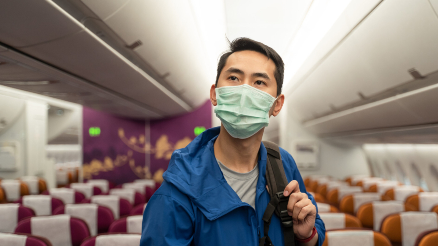 Asian traveler business man with luggage, bag wearing face mask looking overhead storage bin on airplane in airport. Male passenger traveling by plane transportation during covid19 virus pandemic.